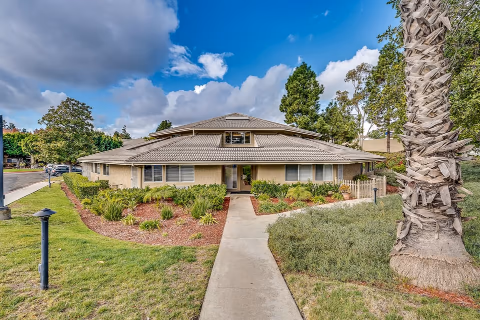 Exterior view of a single-story senior living facility building with a tiled roof, surrounded by landscaped greenery including bushes, trees, and a palm tree. A concrete walkway leads to the entrance under a partly cloudy blue sky.