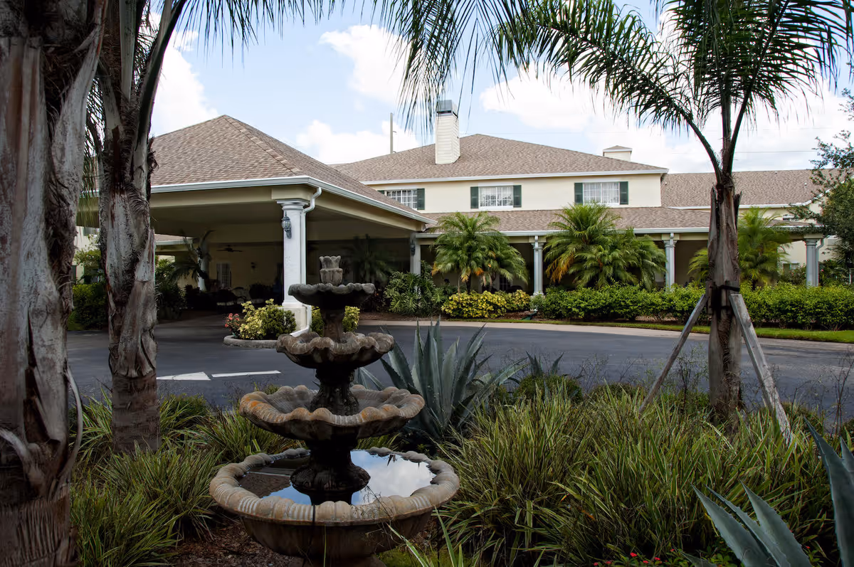View of the exterior of Lexington Manor senior living facility with a covered entrance, palm trees, shrubs, and a three-tiered stone fountain in the foreground under a partly cloudy sky.