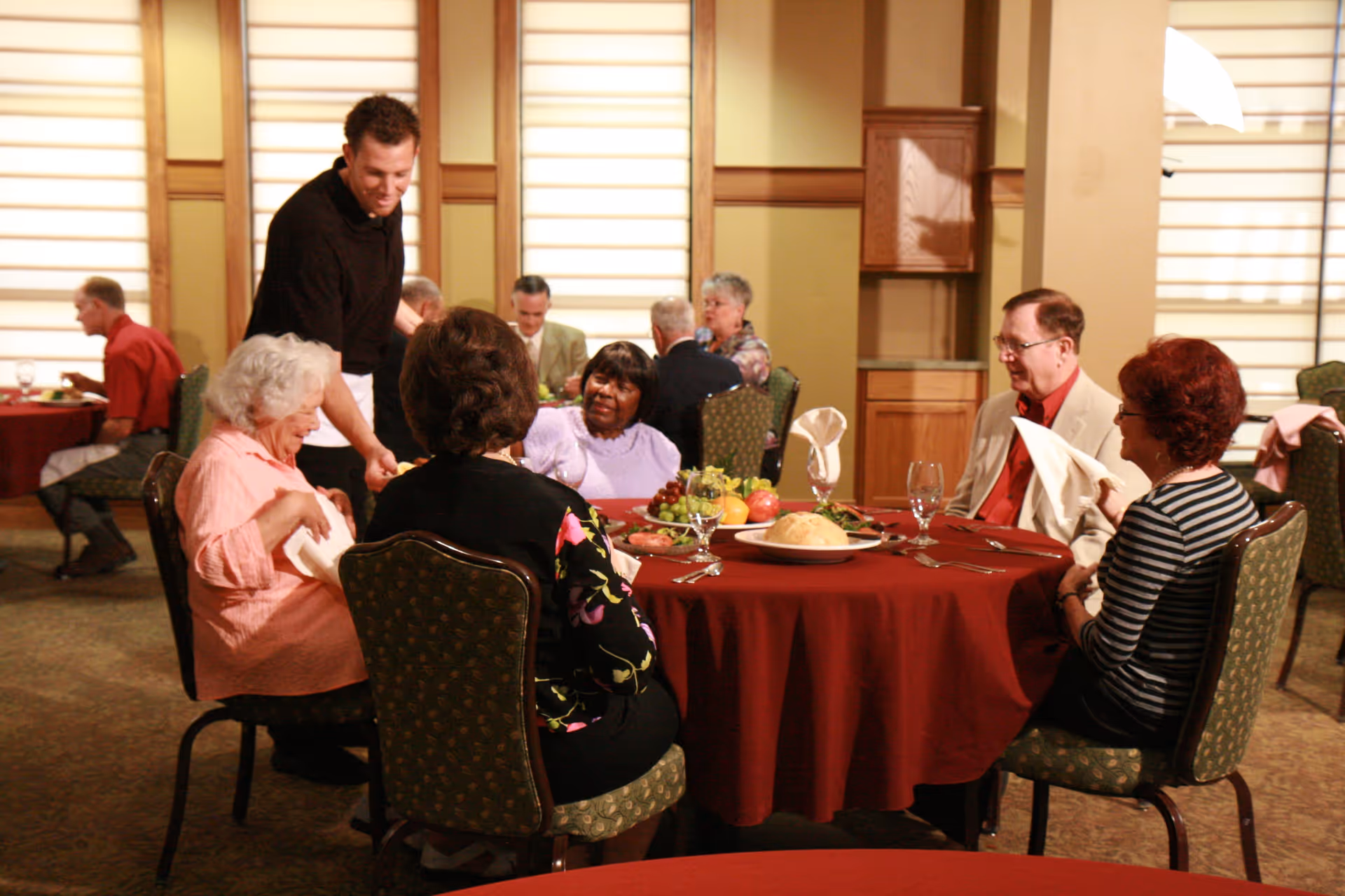 A group of elderly people sitting around a round table with a red tablecloth in a dining room. A waiter is serving them food. The table has plates of fruit, bread, and glasses of water. Other diners are visible in the background.