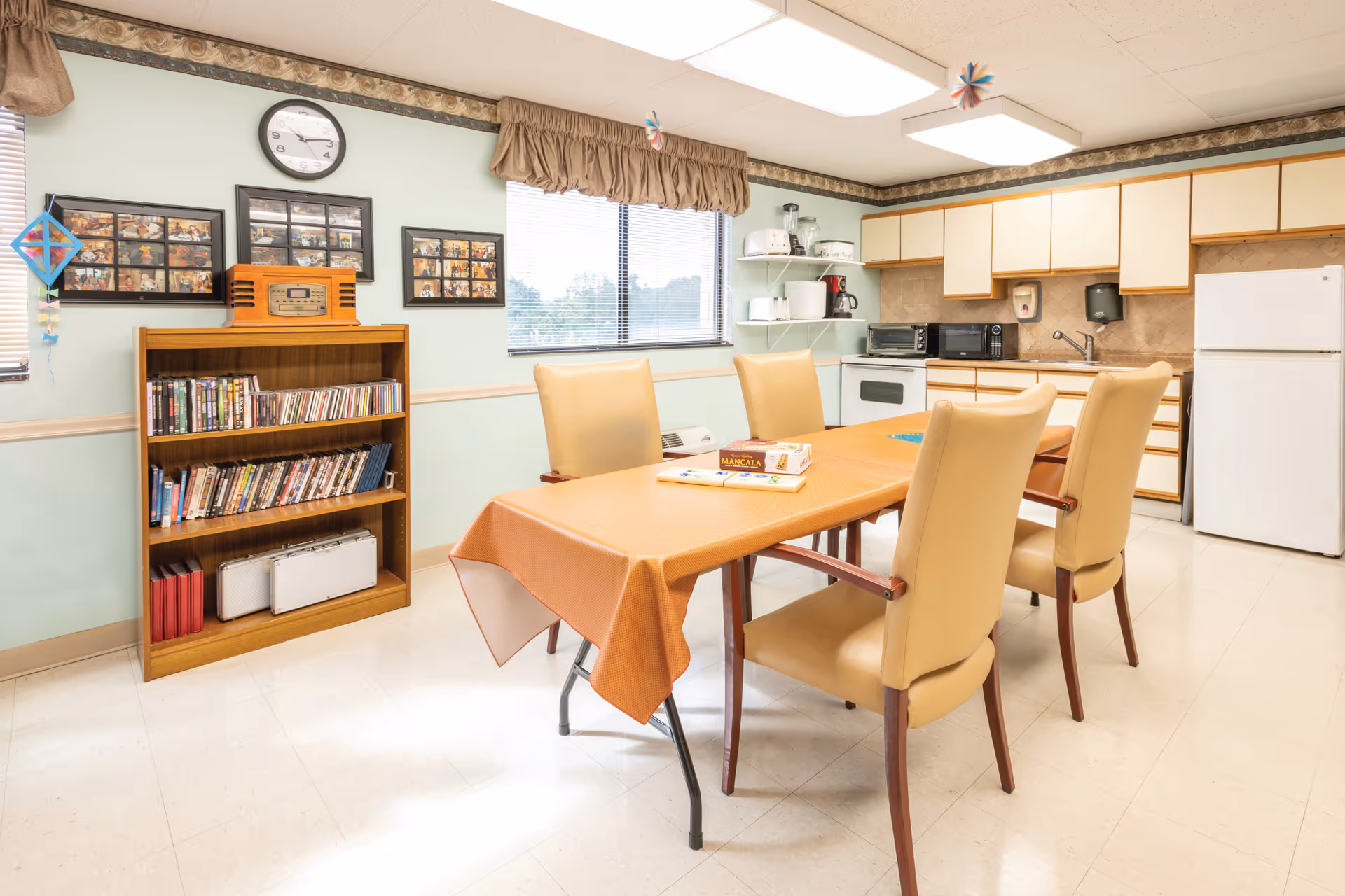 A bright room with a kitchen area and a dining table covered with an orange tablecloth. Four beige cushioned chairs surround the table, which has board games on it. The kitchen has white cabinets, a white refrigerator, a microwave, a toaster oven, and other small appliances. A wooden bookshelf filled with DVDs and books is against the wall, with framed photos and a clock above it. A window with a brown valance lets in natural light.