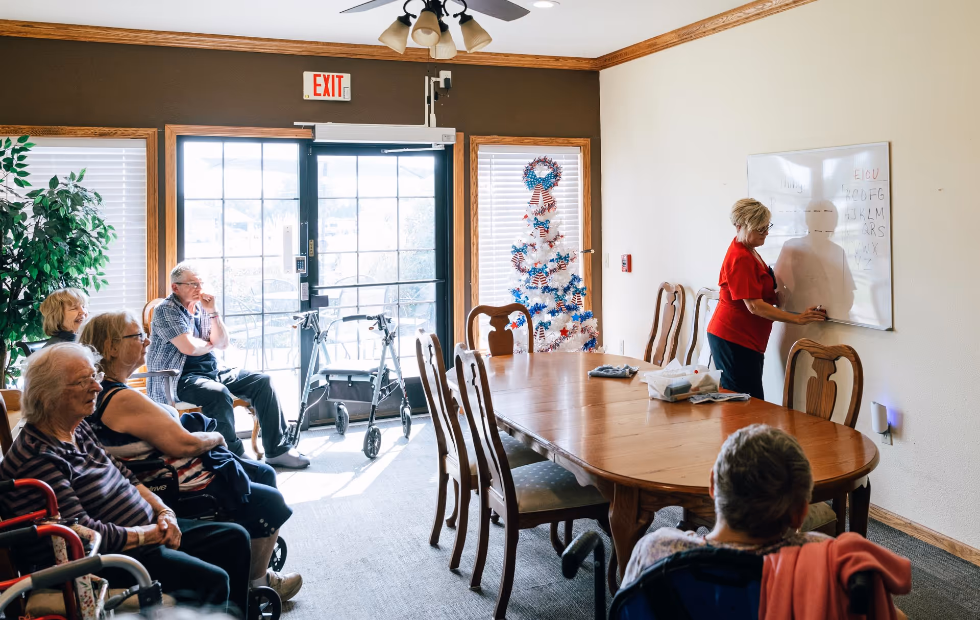 A group of elderly people seated around a large wooden table in a well-lit room with a woman standing at a whiteboard writing. The room has large windows with blinds, a decorated white Christmas tree with red, white, and blue ornaments, and a ceiling fan with lights. Some mobility aids like walkers are visible near the door.