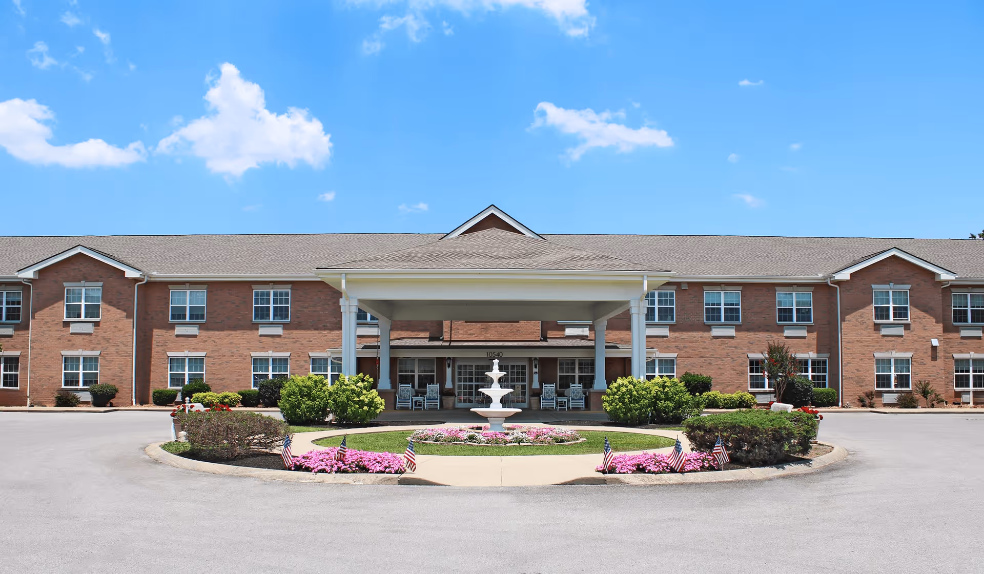 Front exterior view of a two-story brick senior living facility with a covered entrance, a circular driveway, a white fountain in the center, and landscaped bushes and flowers with small American flags.