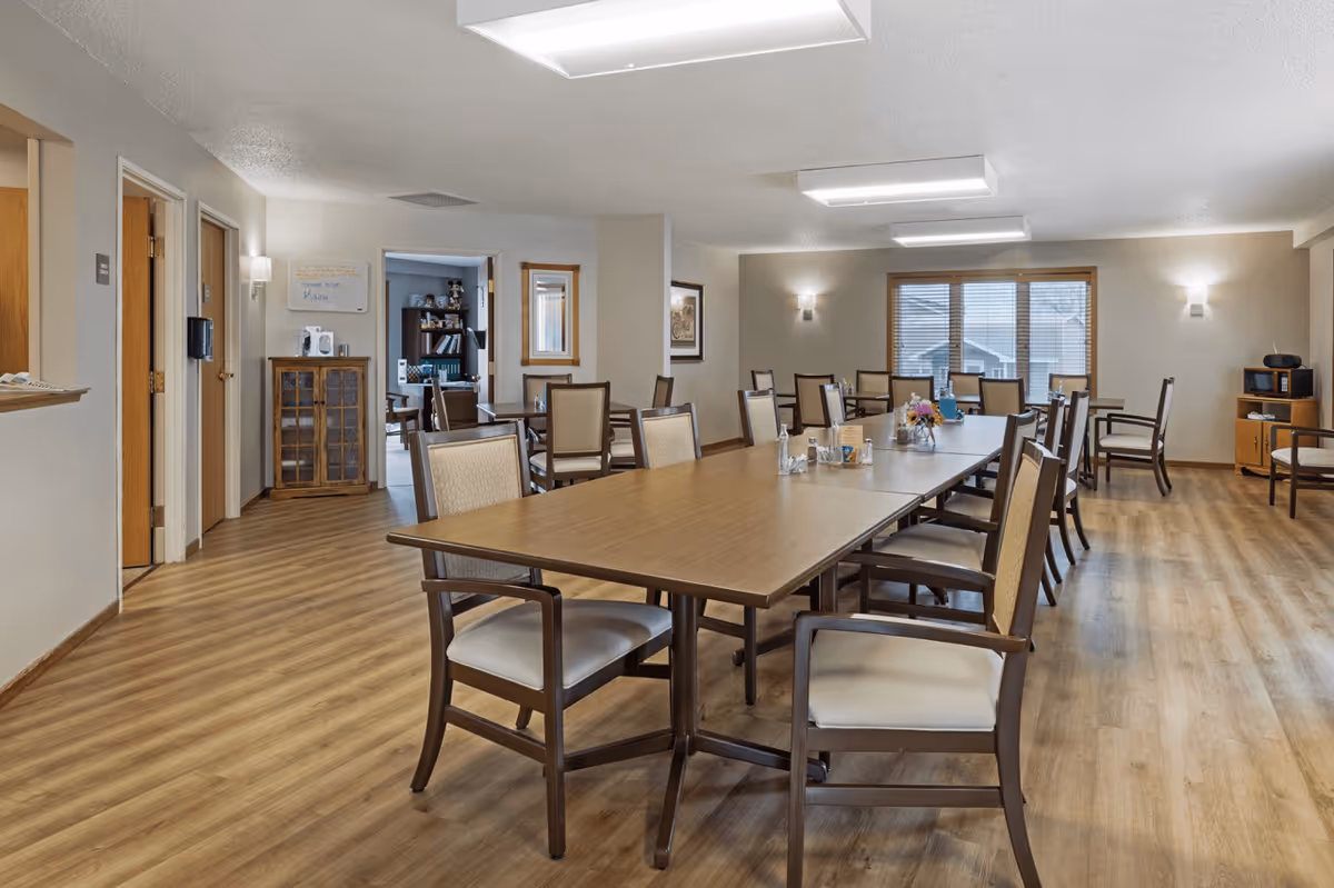 A spacious dining room with wooden floors and several rectangular tables surrounded by cushioned chairs. The room is well-lit with ceiling lights and wall sconces. There are windows with blinds on the far wall, and a small cabinet with a microwave and other items is visible in the corner. The room has a neutral color scheme with beige walls and wooden trim.