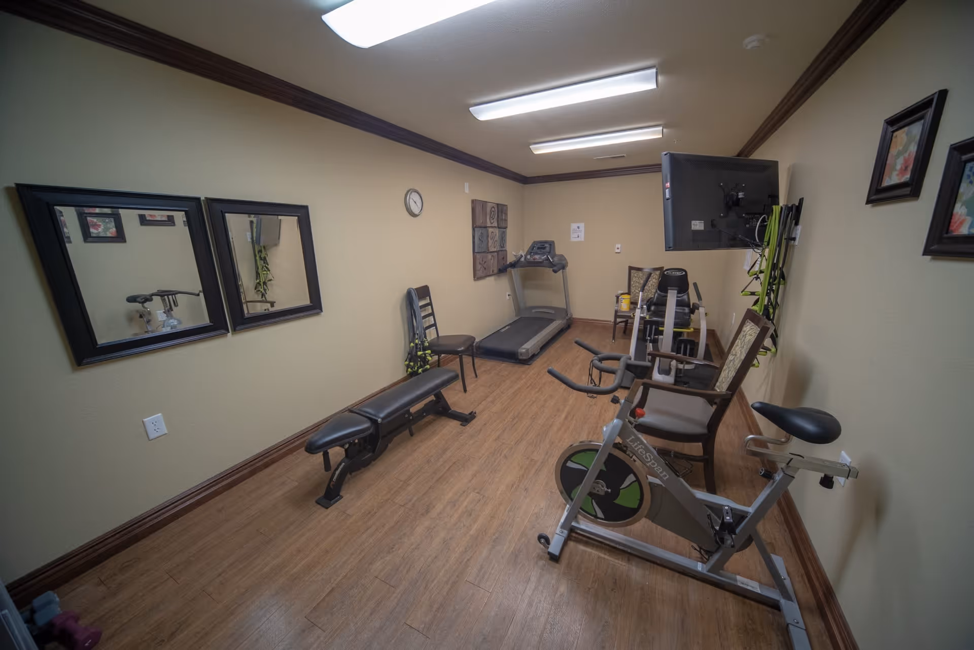 Small fitness room with exercise equipment including a stationary bike, treadmill, and weight bench. The room has wood flooring, beige walls with dark brown trim, two mirrors on one wall, framed artwork, and fluorescent ceiling lights.