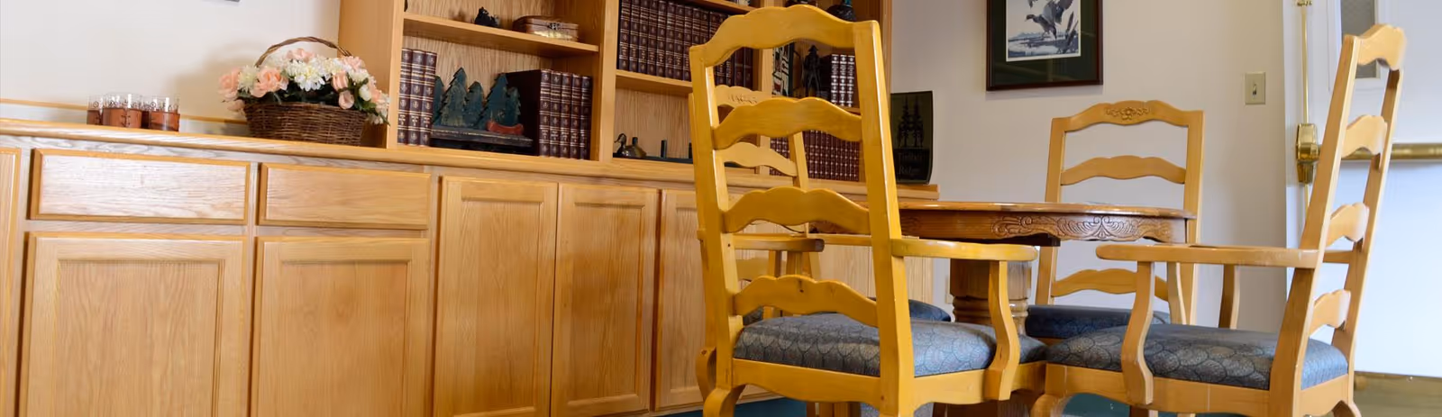 Wooden dining table with matching chairs in front of built-in cabinets and bookshelves holding decorative items and flowers.