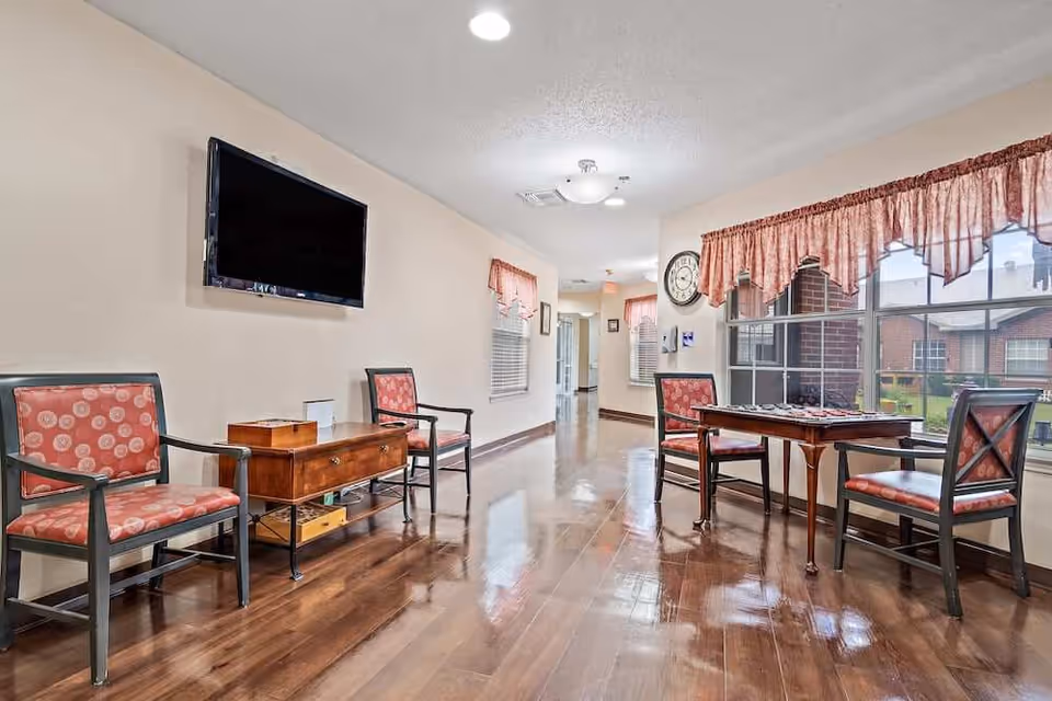 Bright common room with upholstered chairs, a wall-mounted TV, wooden floors, and a small table by large windows.