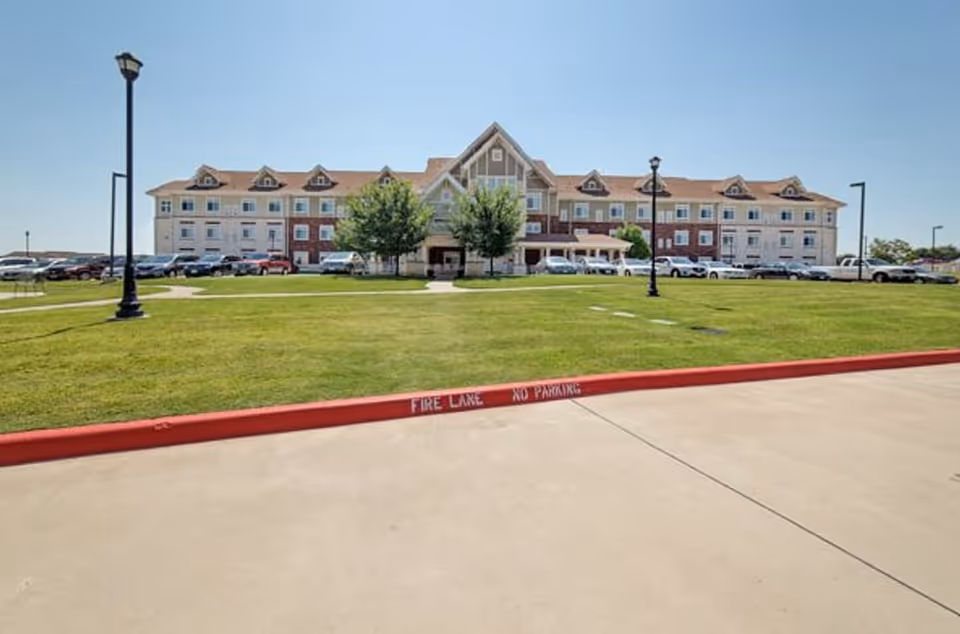 Front exterior view of The Ivy of McKinney, a multi-story senior living facility with a large green lawn and parking area in front under a clear blue sky.