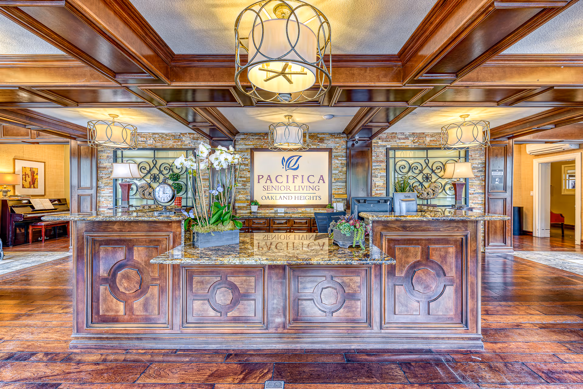 Reception area of Pacifica Senior Living Oakland Heights featuring a wooden front desk with granite countertop, decorative plants, a clock, and lamps. The background has a stone wall with a large sign displaying the facility name. The ceiling has wooden beams and modern light fixtures.