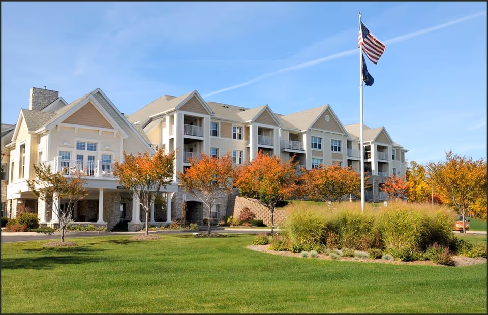Exterior view of a large senior living facility building with beige siding and stone accents, surrounded by green grass and trees with autumn foliage. An American flag and another flag are flying on a flagpole in front of the building under a clear blue sky.