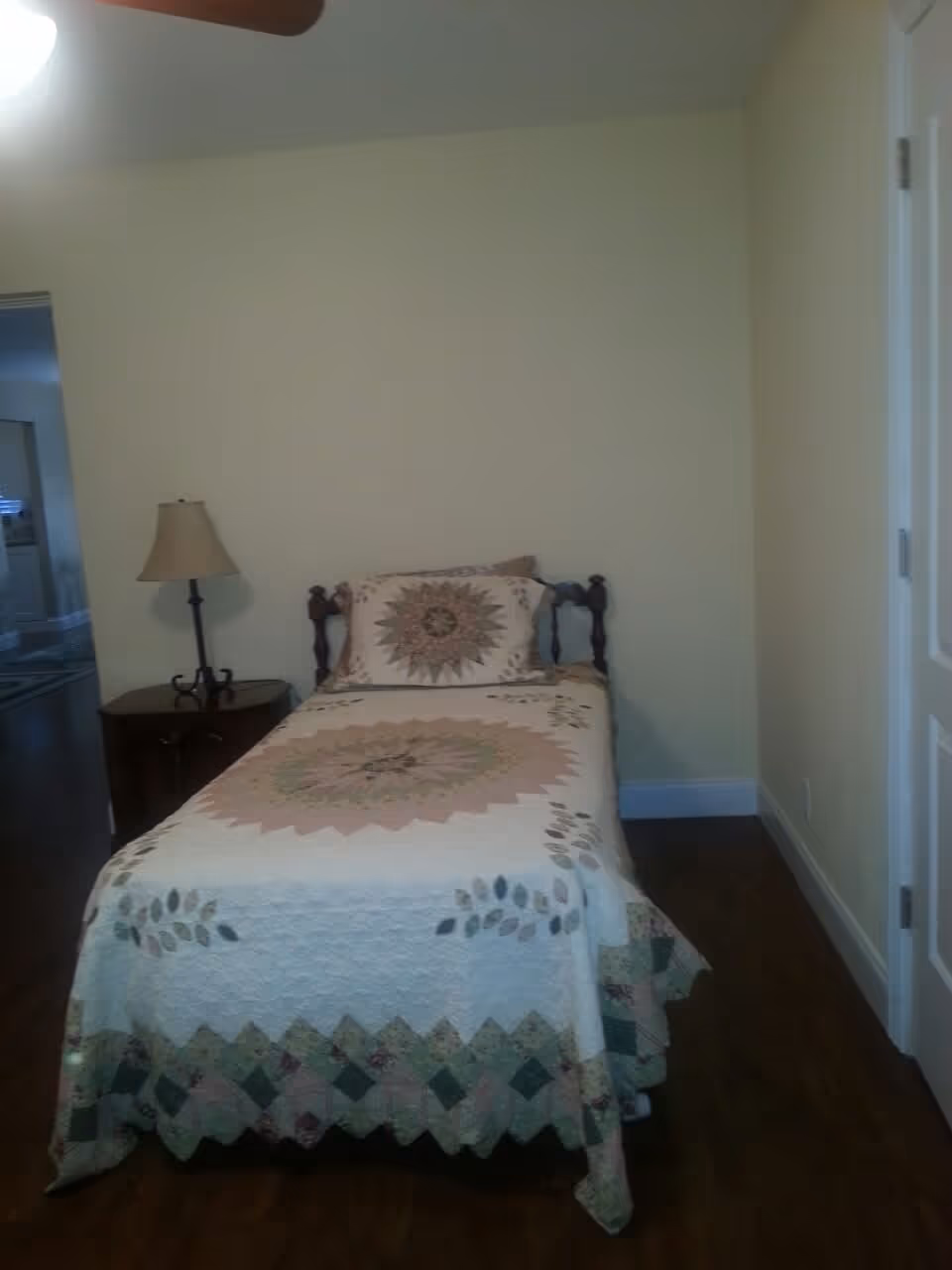 Small bedroom with a single bed covered by a patterned quilt, a nightstand and lamp against a pale wall.