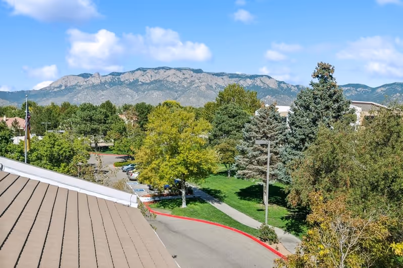 View of a parking lot and green landscaped area with trees and a sidewalk, set against a backdrop of a mountain range under a partly cloudy blue sky.