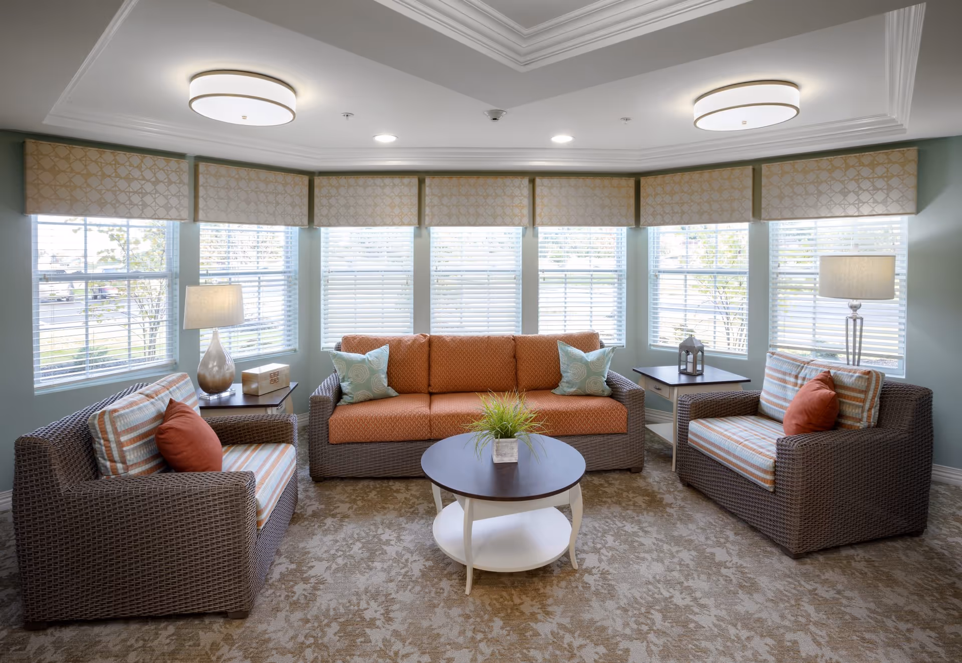 A bright and cozy living room area with a large bay window featuring white blinds and patterned valances. The room contains a wicker-style sofa with orange cushions and two matching armchairs with striped cushions and orange throw pillows. There are two side tables with lamps and decorative items, and a round coffee table with a small green plant in the center. The carpet has a subtle floral pattern, and the ceiling has recessed lighting and two modern round light fixtures.