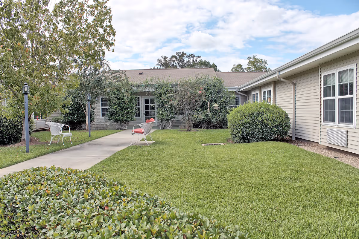 A grassy courtyard with a concrete walkway, metal benches, trimmed shrubs, and single-story building wings under a partly cloudy sky.