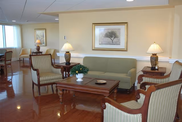 A well-lit living room area with a green sofa, two striped armchairs, a wooden coffee table with a small plant, two side tables with lamps, and framed artwork on the wall. The room has polished wooden floors and additional seating visible in the background near large windows.