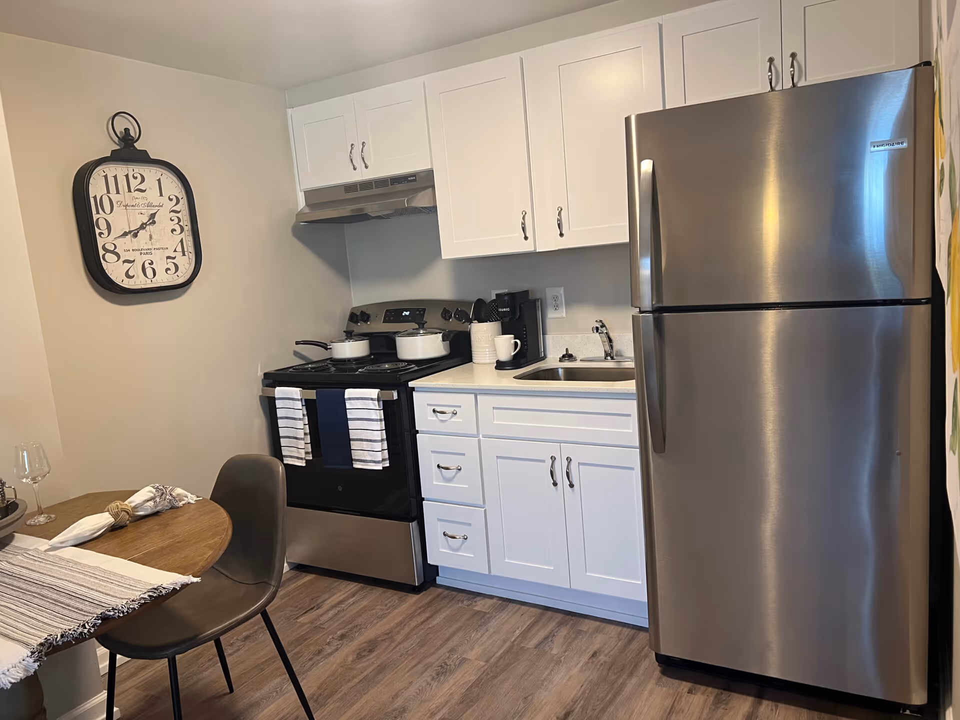 Small modern kitchen with stainless steel refrigerator, stove, white cabinets, sink and a small dining table with a chair.