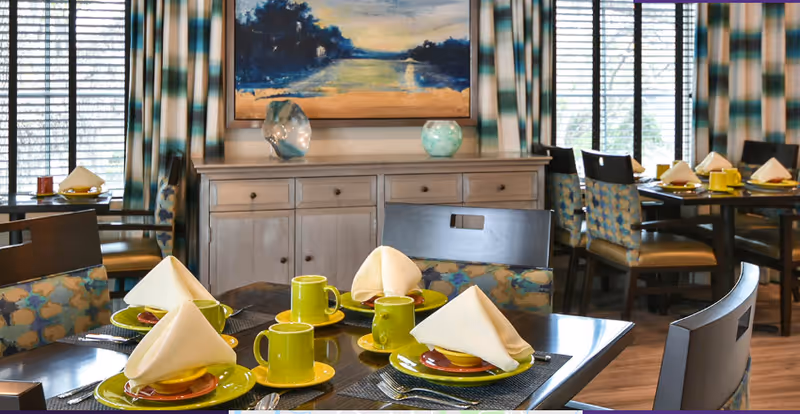 A dining room with multiple tables set for a meal, featuring green cups and yellow plates with folded white napkins. The room has large windows with patterned curtains, wooden chairs with patterned cushions, a wooden sideboard, and a landscape painting on the wall.