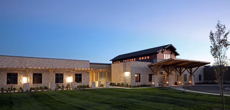 Front exterior of a stone-clad senior living building with a covered entrance, illuminated windows, and a well-kept lawn at dusk.