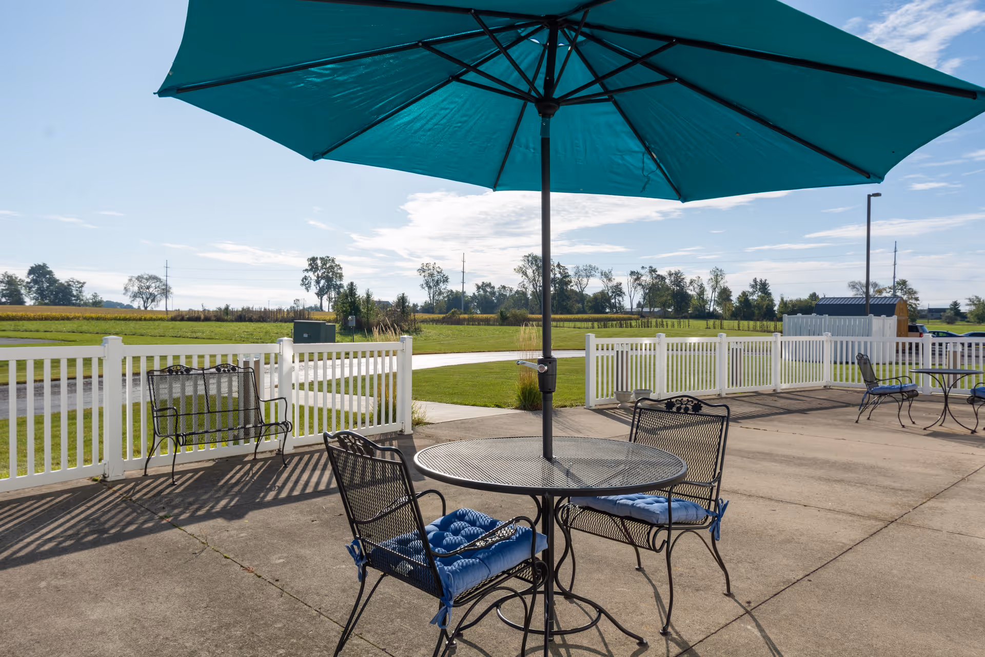 Outdoor patio area with metal tables and chairs, some with blue cushions, under large teal umbrellas. The patio is surrounded by a white picket fence and overlooks a grassy field with trees and a clear sky.