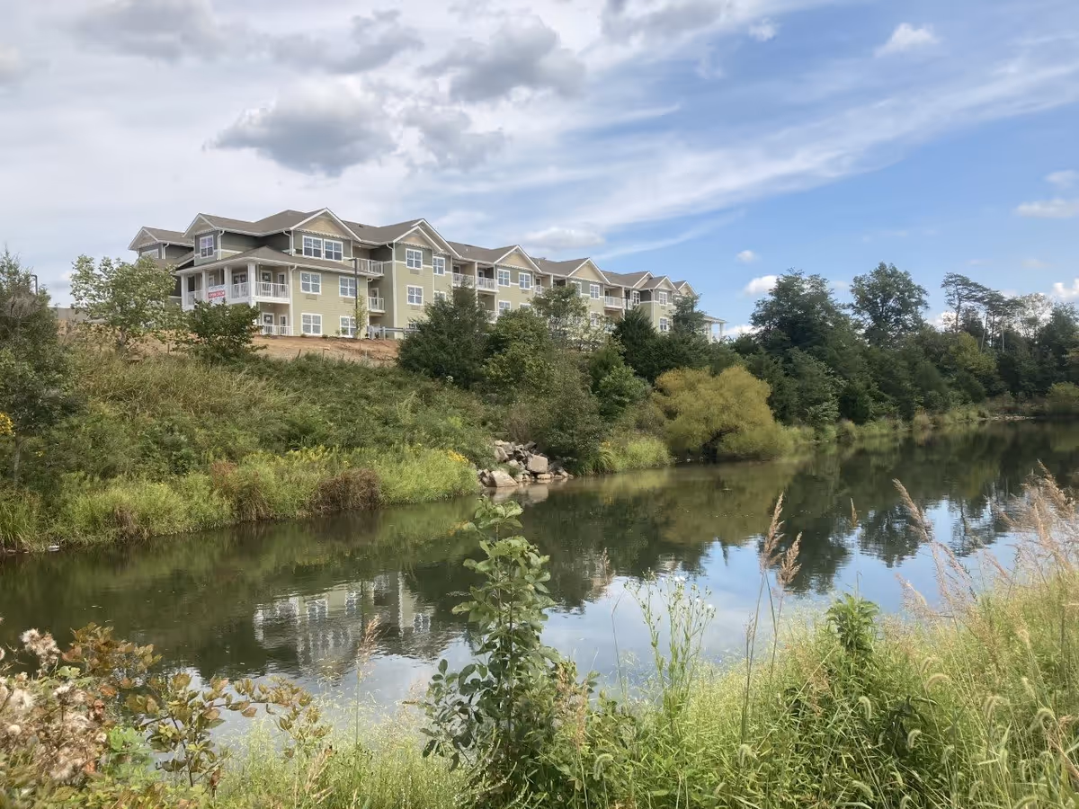 Multi-story pale green senior living building on a hillside reflected in a calm pond with trees and grassy foreground.