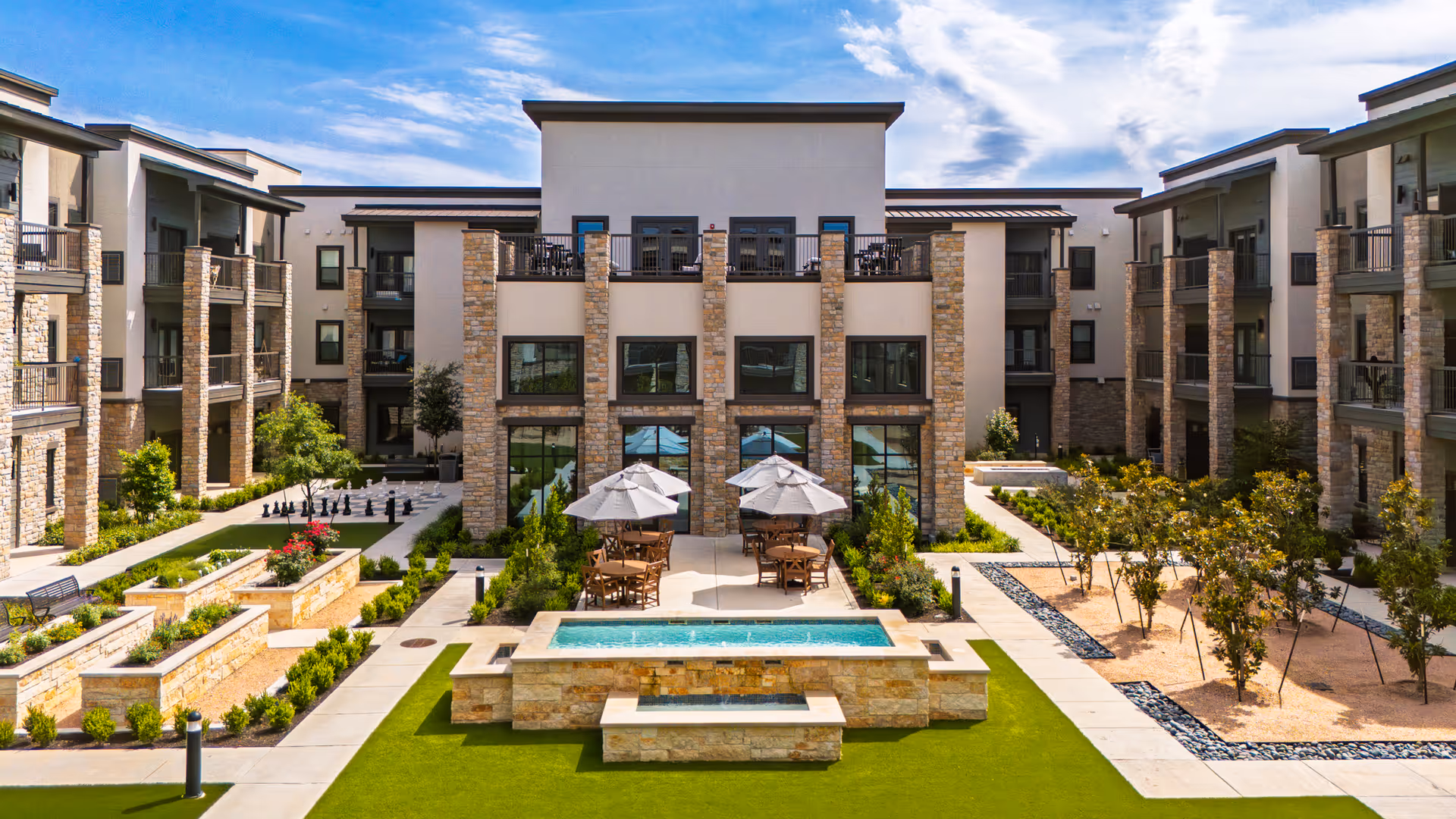Outdoor courtyard area of a senior living facility with a rectangular water feature in the center, surrounded by green artificial turf and stone planters with flowers and shrubs. There are tables with umbrellas and chairs on a paved patio area in front of a modern three-story building with stone and stucco exterior. The sky is partly cloudy.