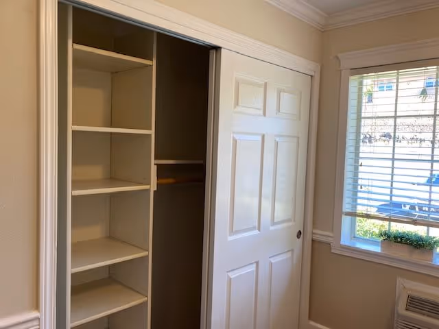 Interior view of a room with a white sliding closet door partially open, revealing empty shelves and a hanging rod inside. To the right, there is a window with white blinds and a small green plant on the windowsill. The walls are painted beige with white trim.