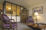 Cozy furnished living room with two upholstered armchairs, a coffee table, lamp, and a glass-front cabinet.