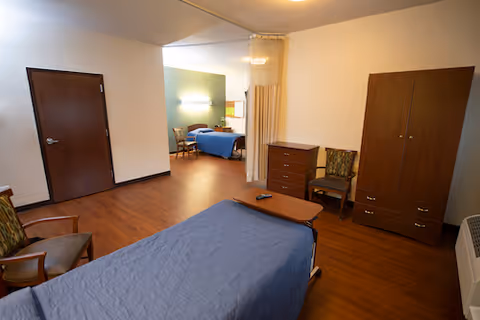 A spacious senior living bedroom with two beds, wooden furniture, chairs, and a privacy curtain dividing the area.
