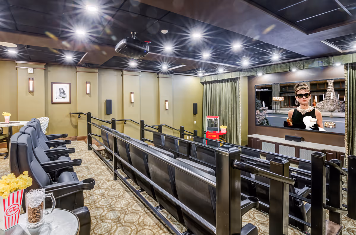 A small private movie theater room with two rows of black leather reclining seats, a popcorn machine, and a large screen showing a scene from the movie Breakfast at Tiffany's. The room has beige walls with framed pictures, wall sconces, and a patterned carpet.