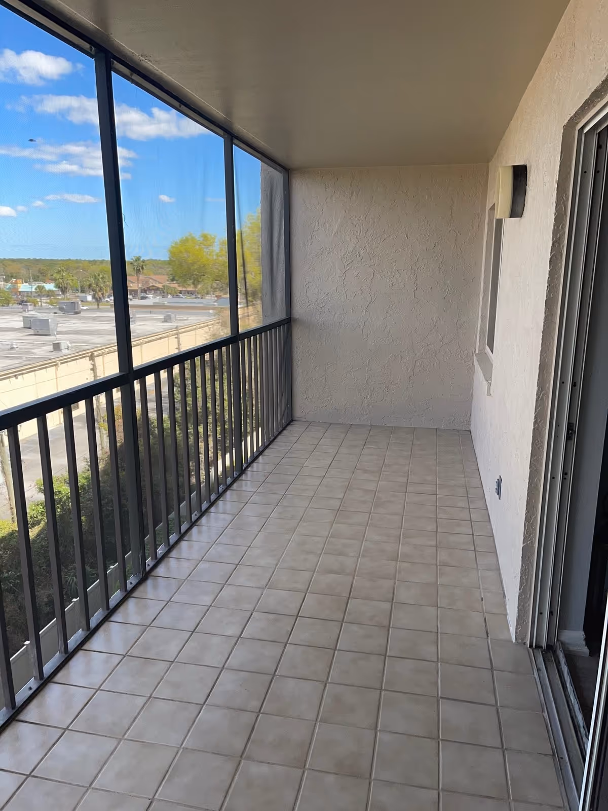 A tiled balcony with a metal railing and screened enclosure. The balcony overlooks a parking lot and some trees under a blue sky with scattered clouds. The balcony has a textured white wall and a sliding glass door leading inside.
