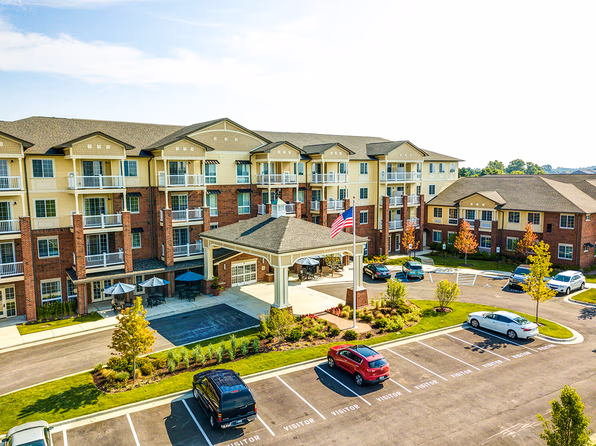 Exterior view of Independence Village of Zionsville West, a multi-story senior living facility with a covered entrance, balconies, and a parking lot with several cars. The building features a combination of brick and light-colored siding with landscaped greenery and an American flag near the entrance.