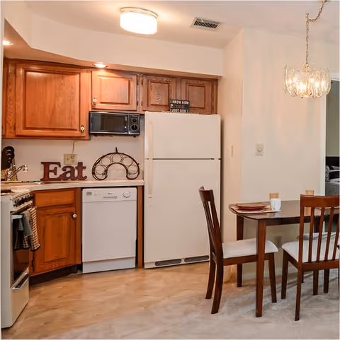 Kitchen area with oak cabinets, a white refrigerator and dishwasher beside a small dining table with two chairs.