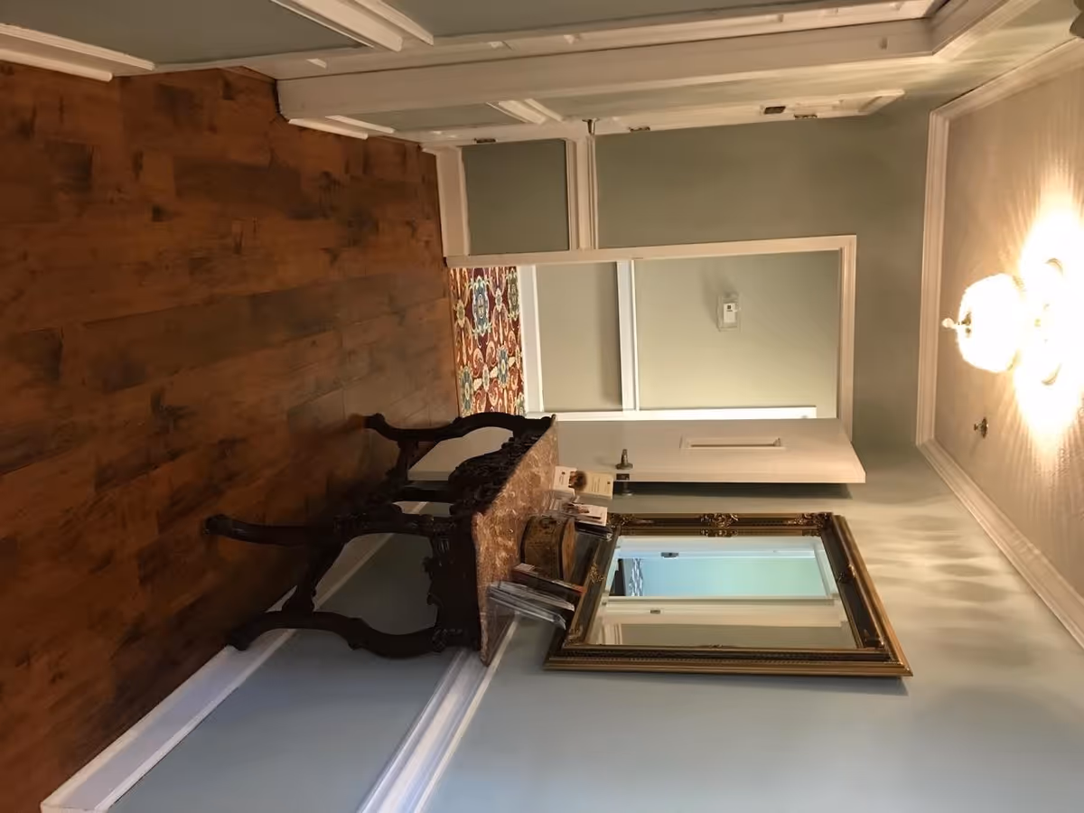 A hallway with wooden flooring and light blue walls featuring white trim. There is an ornate wooden table with a marble top against the wall, holding brochures and a decorative box. Above the table is a large, gold-framed mirror. A white door is open at the end of the hallway, revealing a colorful patterned rug in the adjoining room. A ceiling light fixture is visible on the right side of the image.