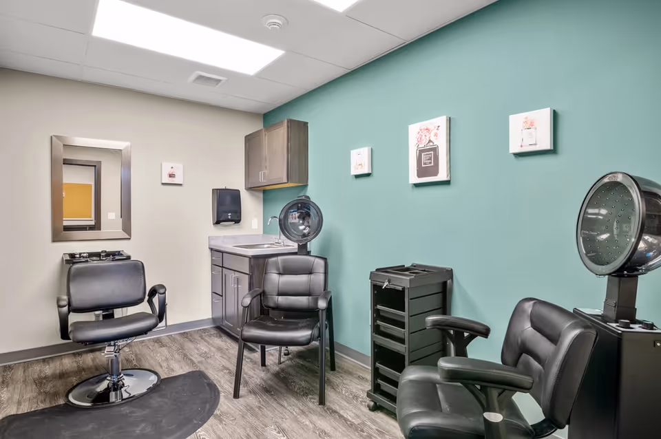Salon room with black styling chairs, hooded hair dryers, a sink and cabinets against a teal accent wall with artwork.