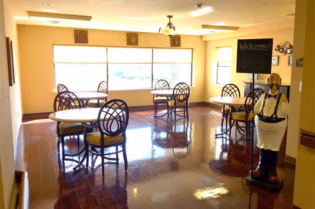 A bright dining room with several round tables and chairs arranged neatly. Large windows allow natural light to fill the room. A decorative statue of a waiter holding a chalkboard sign that says 'Welcome!' stands near the entrance.