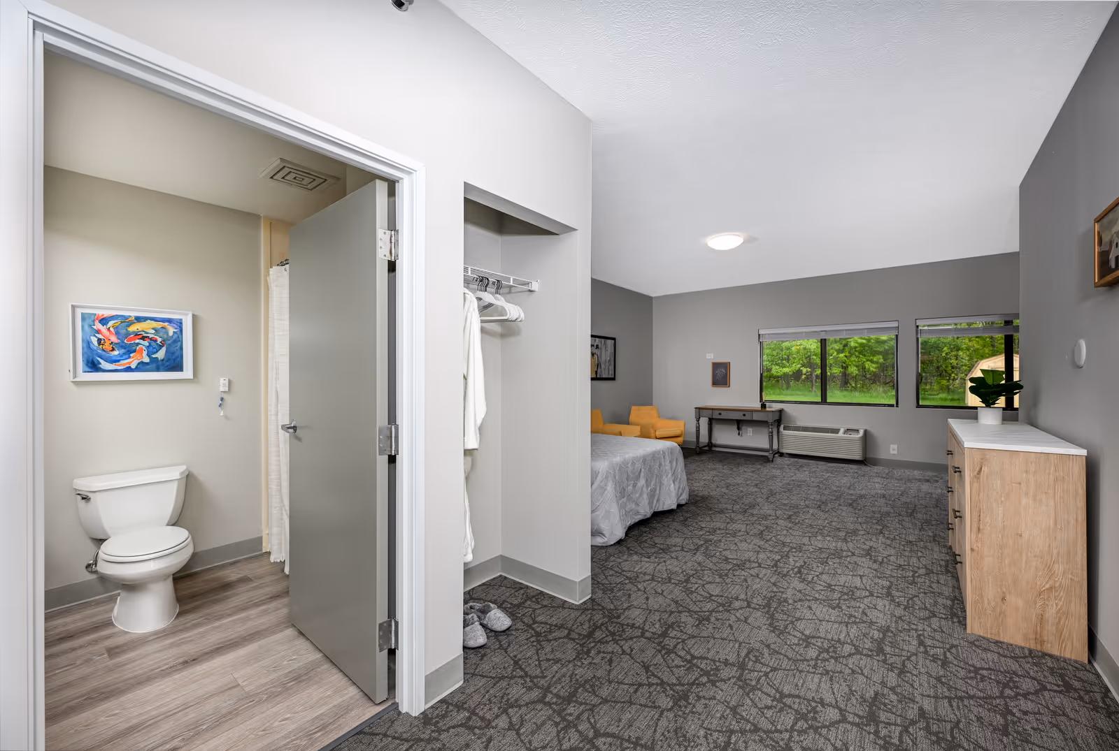 A spacious senior living facility room with a gray carpet and walls. The room features a bed with a gray bedspread, a yellow armchair, a desk under two large windows showing green trees outside, and a wooden dresser with a plant on top. To the left, there is an open door leading to a bathroom with a toilet, a shower curtain, and a framed picture of colorful koi fish on the wall. There is also an open closet space with hanging robes and slippers.