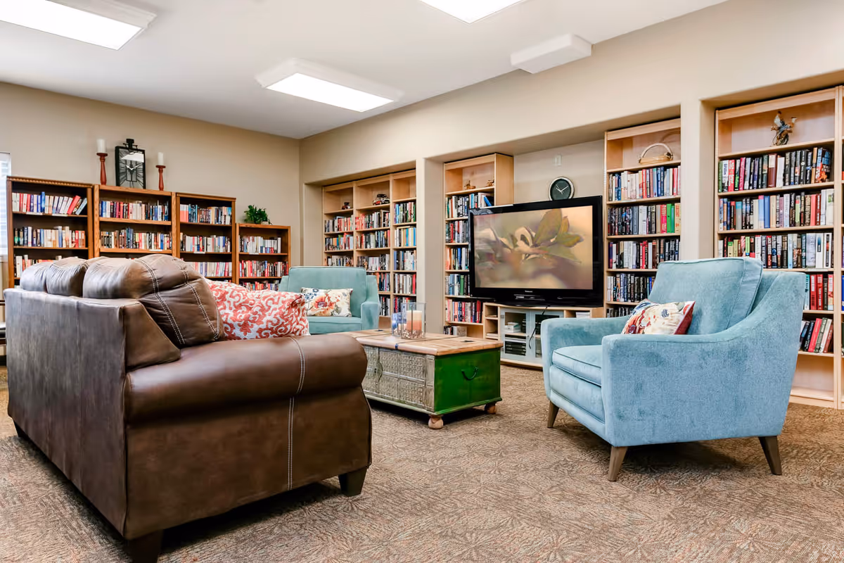 A cozy living room area with a brown leather sofa, two blue armchairs with patterned cushions, a wooden coffee table with a green base, and multiple bookshelves filled with books. A flat-screen TV is mounted on a stand between the bookshelves, displaying an image of a bird. The room has beige walls and carpeted flooring.