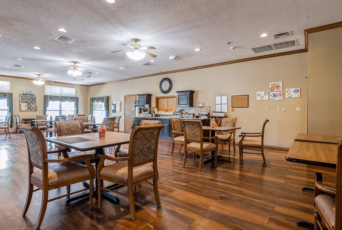 A spacious dining room with wooden floors and multiple tables and chairs arranged neatly. The room has ceiling fans with lights, large windows with green valances, and a wall clock above a counter area with cabinets and a coffee machine. Artwork and bulletin boards are displayed on the walls.
