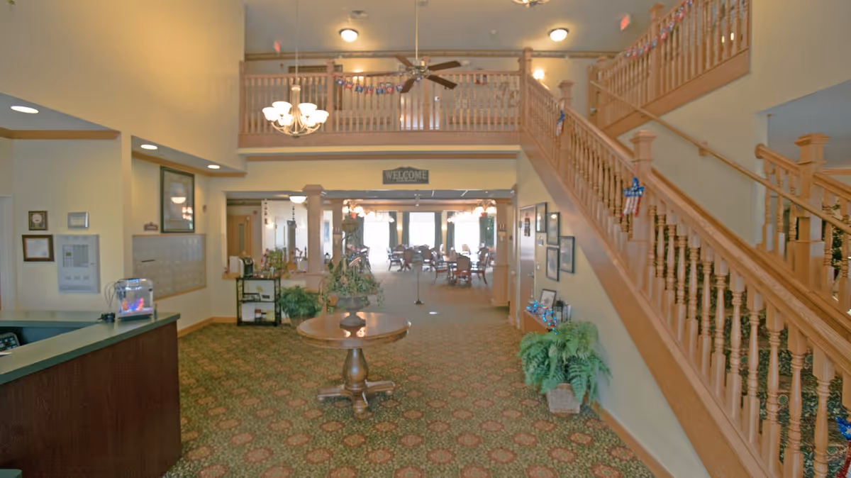 Interior view of a senior living community lobby with a wooden staircase on the right, a round wooden table with a plant in the center, a reception desk on the left, and a dining area with tables and chairs visible in the background. The space is decorated with plants, framed pictures, and patriotic decorations.