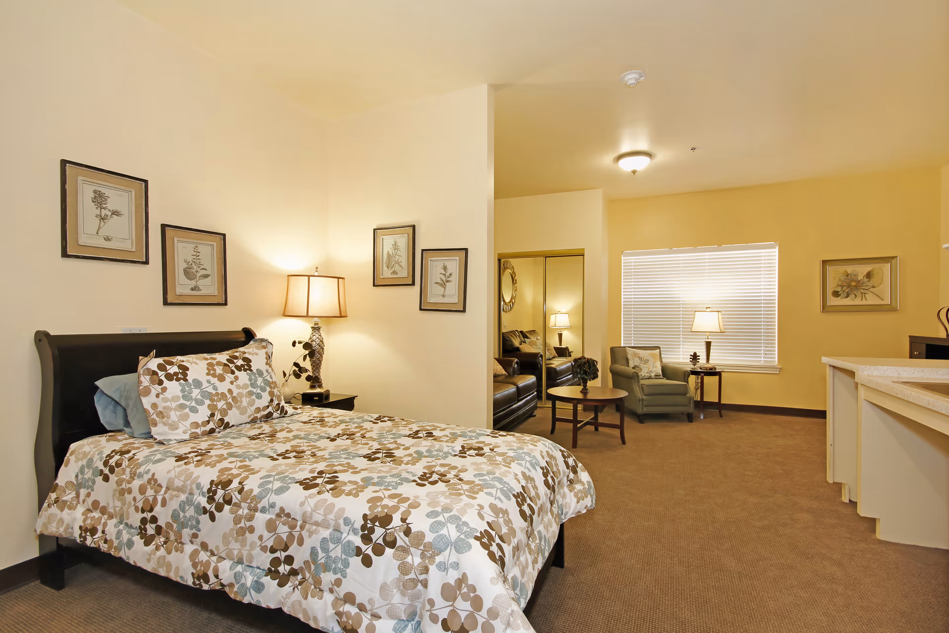 A cozy senior living facility room at Sodalis Wharton featuring a bed with floral patterned bedding, a bedside table with a lamp, framed botanical prints on the wall, a sitting area with a green armchair, a round coffee table, a sofa, and a window with blinds. The room has beige walls and carpeted flooring.