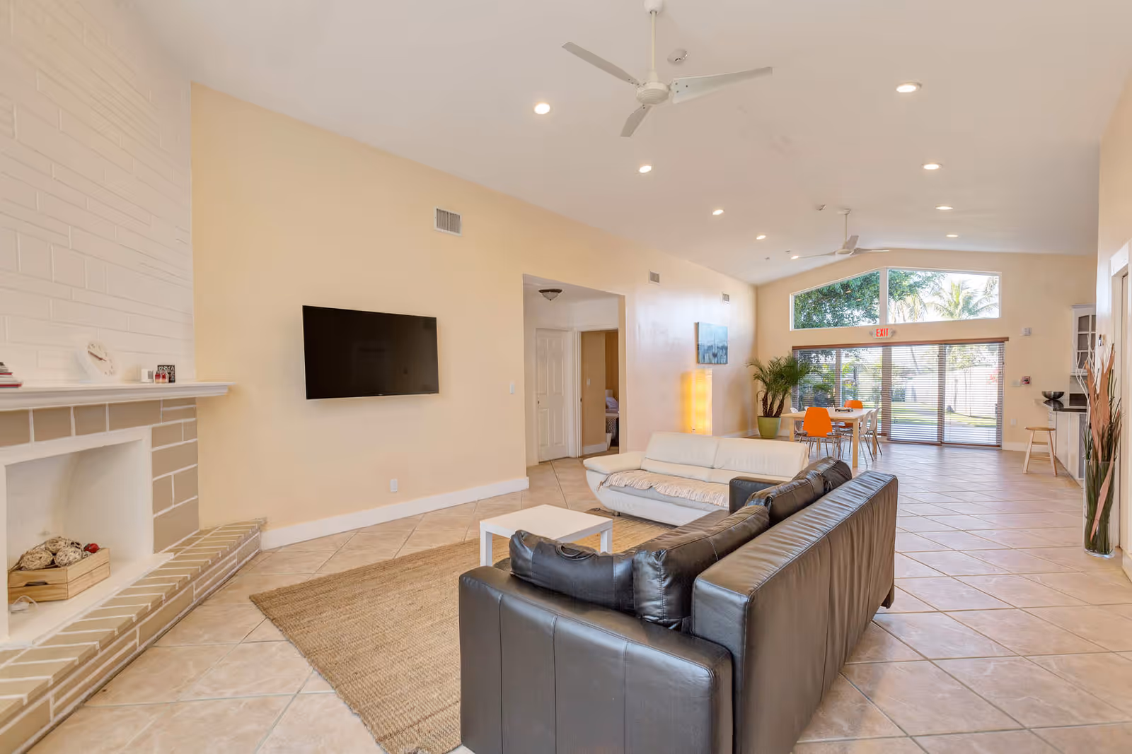 A spacious living room area in an assisted living facility with beige walls and tiled floors. The room features a white couch and a black leather couch arranged around a small white coffee table on a woven rug. A flat-screen TV is mounted on the wall above a beige brick fireplace. In the background, there is a dining area with a table and orange chairs near large windows and glass doors that let in natural light. Ceiling fans and recessed lighting are visible on the ceiling.