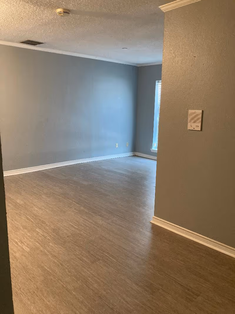 Empty room with light brown wood flooring and gray walls, featuring a window with blinds on the far wall and a smoke detector on the ceiling.