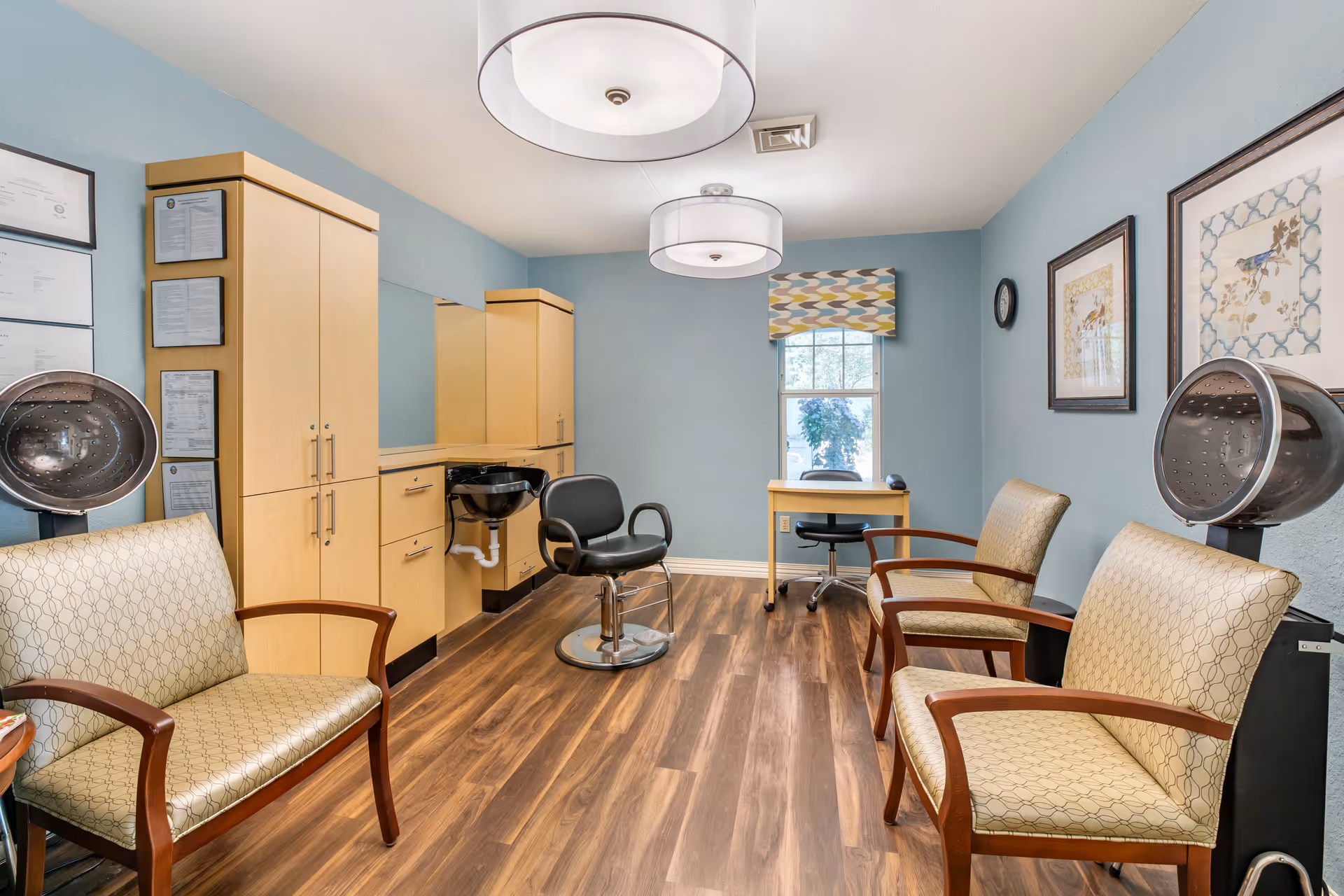 A well-lit hair salon room with light blue walls and wood flooring. The room features two hair drying chairs with hooded dryers, a black salon chair in front of a sink, light wood cabinetry, a small desk with a rolling chair near a window, and three upholstered armchairs with wooden arms. The walls are decorated with framed certificates and artwork.