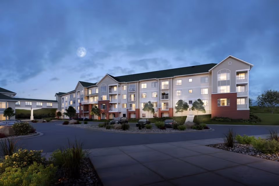 Exterior view of a multi-story senior living facility building at dusk with lights on inside. The building has a combination of white and red brick facade with balconies and large windows. There is a driveway and landscaped greenery in front, with a partly cloudy sky and a visible moon.