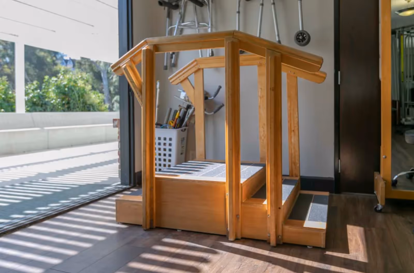 A wooden rehabilitation step platform with handrails on both sides, placed indoors near a large open door leading outside. The platform has three steps with non-slip surfaces. In the background, there is a basket with various tools and equipment, and a mirror on the right side.