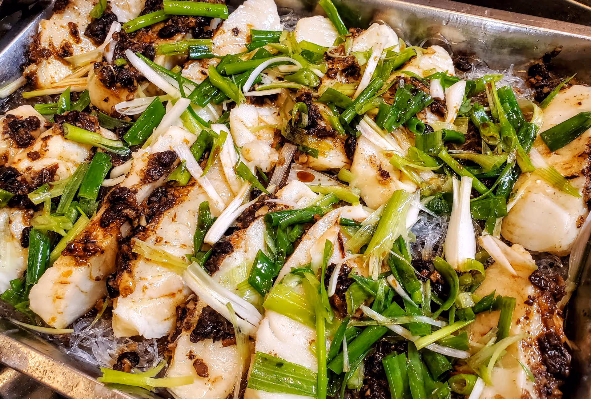 Close-up of a tray filled with cooked white fish fillets garnished with chopped green onions and a dark seasoning or sauce.