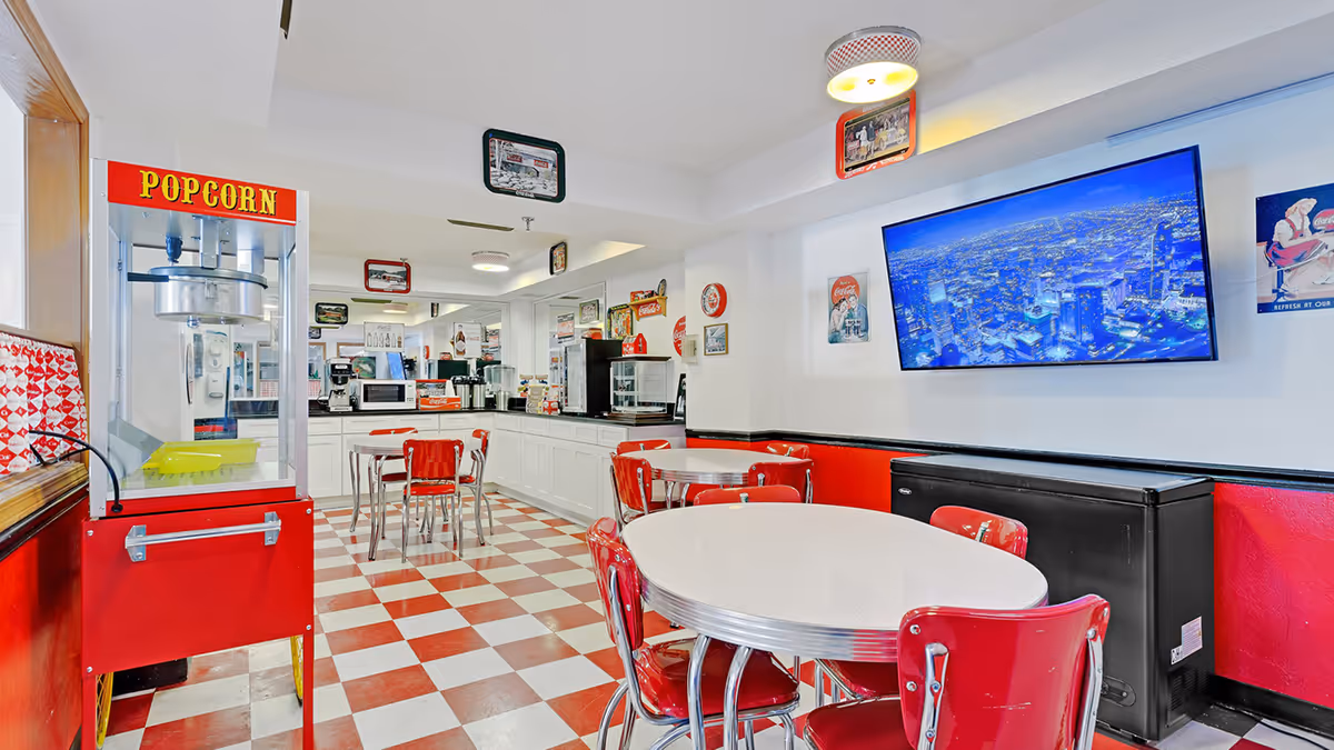 Retro-style communal dining area with red-and-white checkered floor, round tables and chairs, a popcorn machine, and a wall-mounted TV.