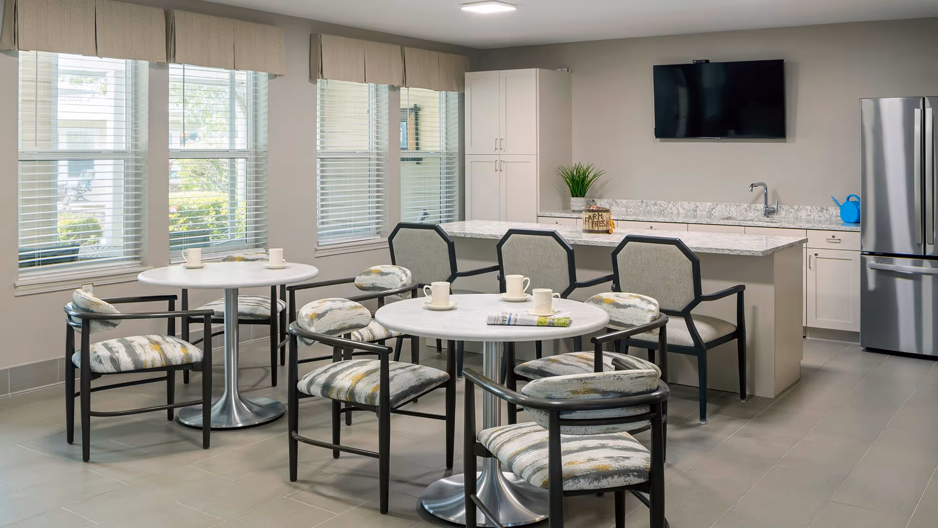 A bright and modern dining area with two round tables surrounded by cushioned chairs featuring patterned upholstery. In the background, there is a kitchen island with a granite countertop, three chairs, a sink, white cabinets, a stainless steel refrigerator, and a wall-mounted flat-screen TV. Large windows with blinds allow natural light to fill the room.