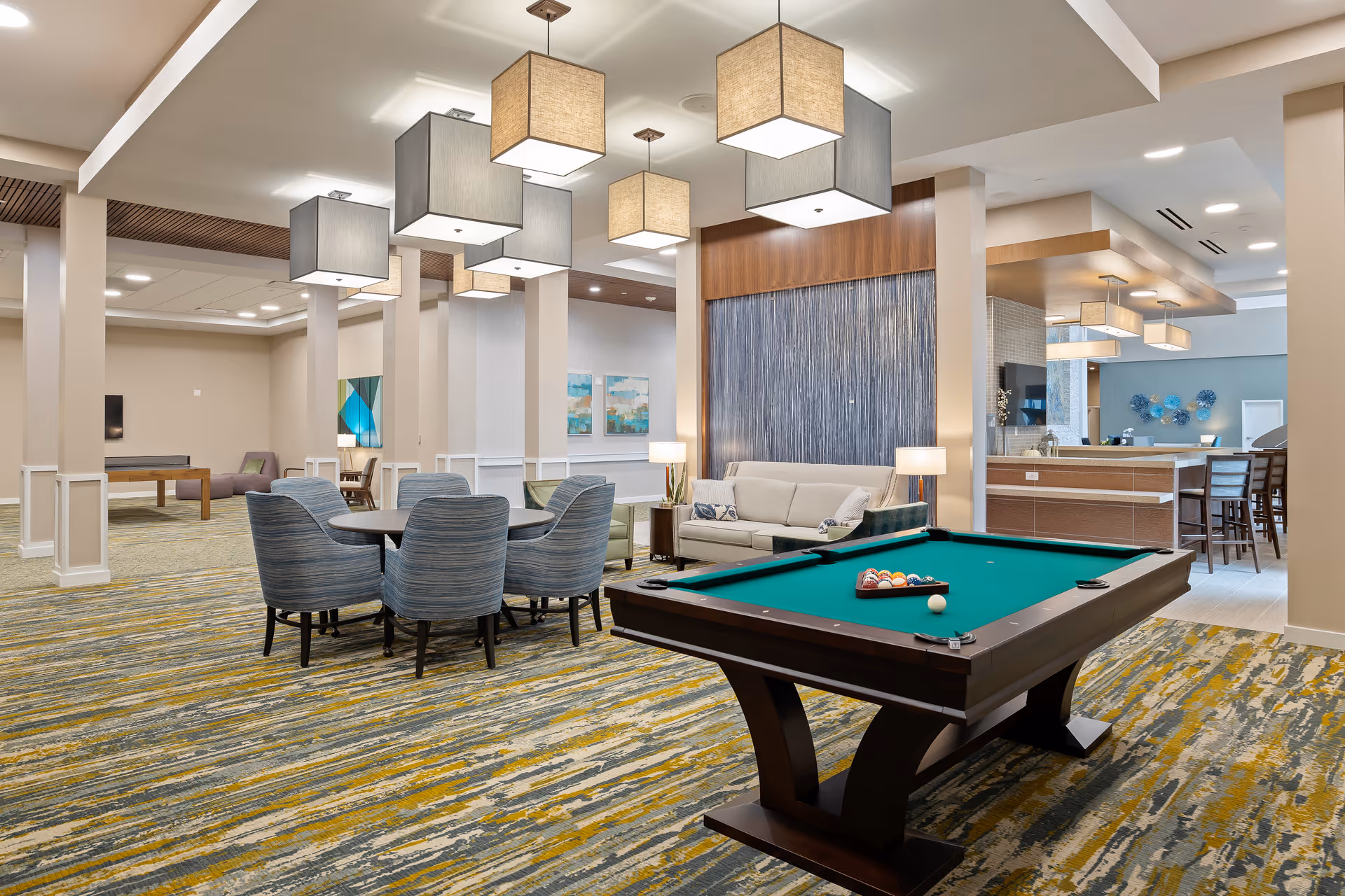 A spacious and well-lit common area in a senior living facility featuring a pool table with balls arranged for a game, a round table with six upholstered chairs, a beige sofa with side tables and lamps, modern pendant lights hanging from the ceiling, and a bar area with high chairs in the background. The room has a patterned carpet and contemporary wall art.