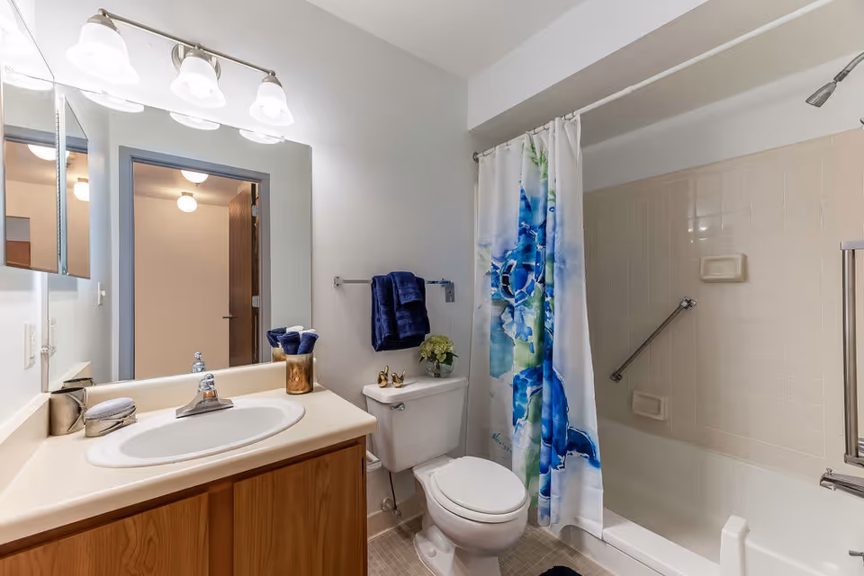 A clean bathroom with a white sink and wooden cabinet, a large mirror with three light fixtures above it, a toilet with a blue towel hanging on the wall above it, and a bathtub with a colorful blue and green shower curtain featuring a sea turtle design.