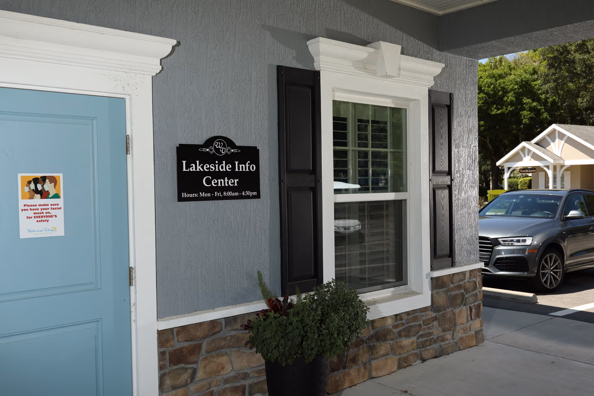 Exterior view of the Lakeside Info Center building with a blue door featuring a sign reminding visitors to wear masks for safety. The building has gray walls with stone accents and a window with black shutters. A black planter with green plants is placed near the window. A parked gray SUV and another small building labeled 'Trolley' are visible in the background.